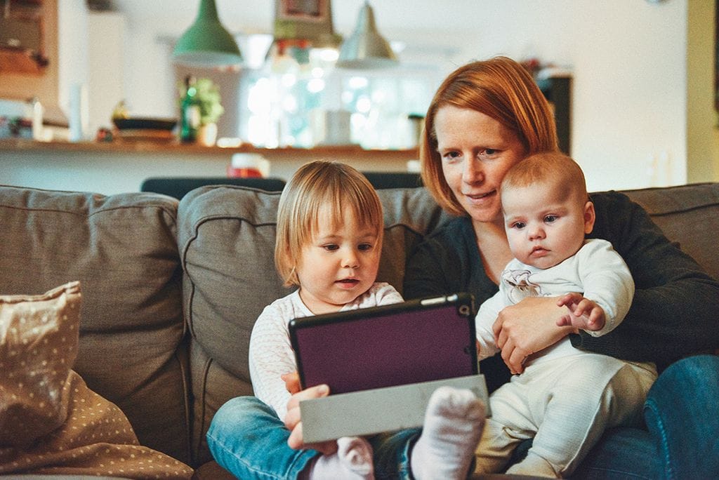 An infant, toddler, and woman sitting on a couch looking at a tablet 